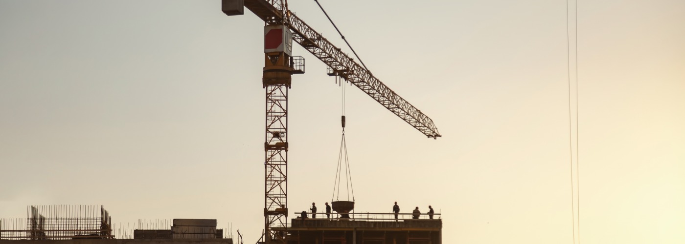 Workers are visible at a construction site, engaged in various tasks with cranes lifting materials. The sky shifts to warmer tones as the sun sets, creating a dynamic atmosphere.